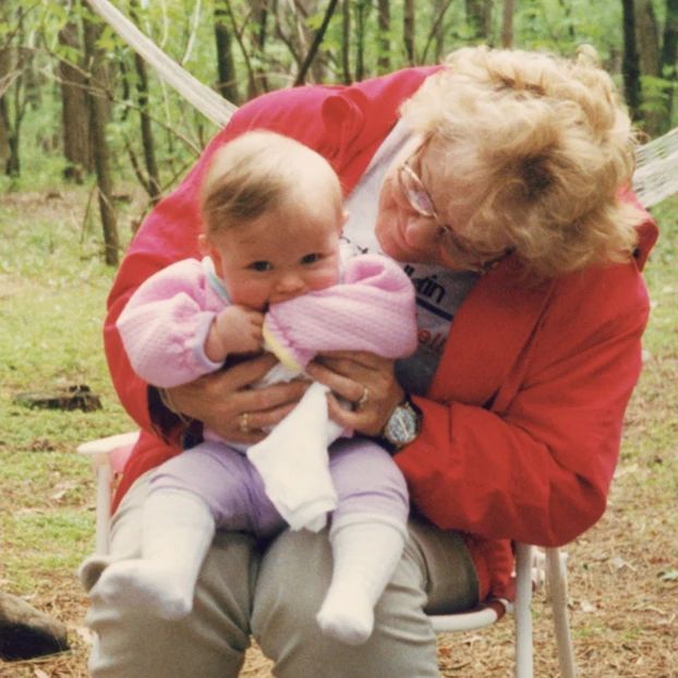 Grandma Kathy holding a baby (me) on her lap while we are camping outdoors. Both of us are smiling.